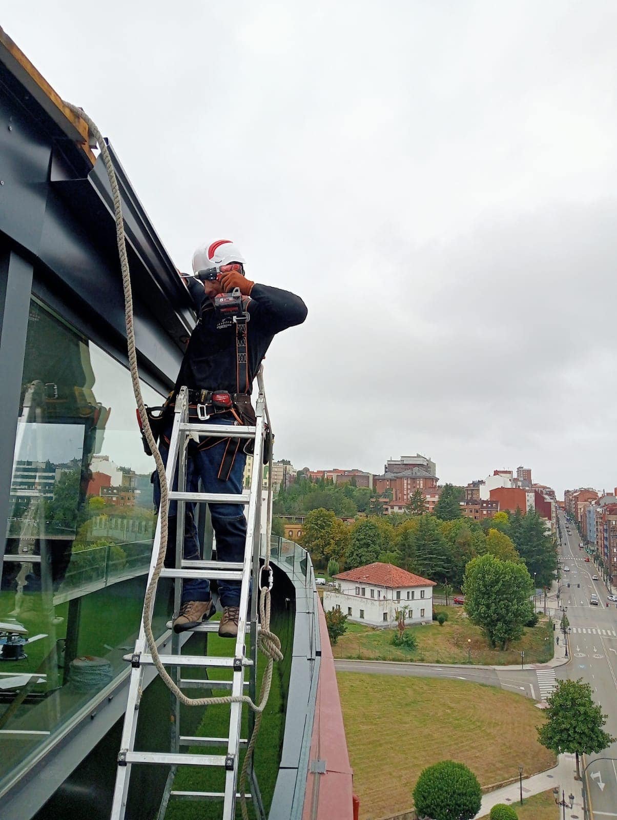 Instalador de ALCUZINC con casco y arnés montando un canalón de aluminio negro en la cubierta de un ático, con la ciudad al fondo bajo un cielo nublado